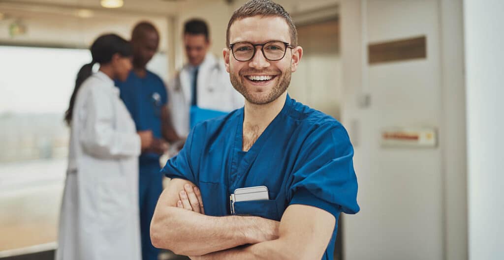 Smiling optimistic young surgeon standing in front of his colleagues with folded arms