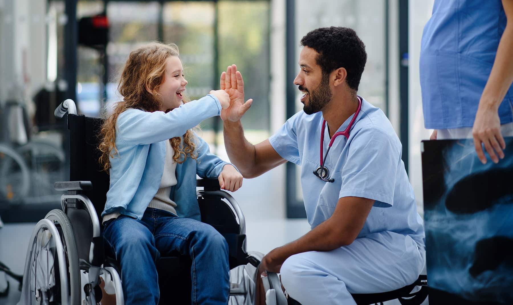 Young doctor having fun with little girl on a wheelchair.